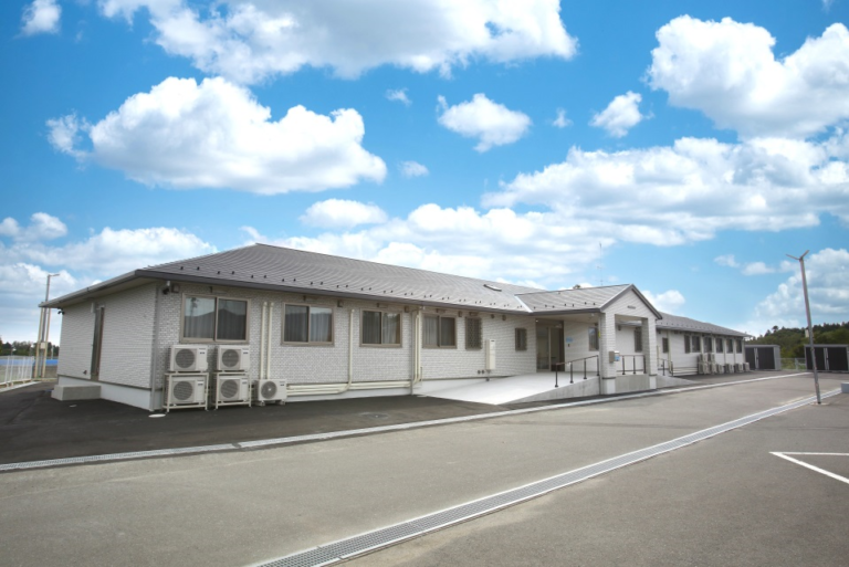 介護施設の白い平屋建物外観。駐車場から見た日中の全景写真。